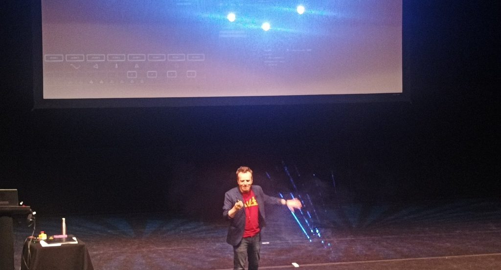 Seb Lee-Delisle presenting on stage, with lasers pointed against the back screen and shining through haze created near his arms.