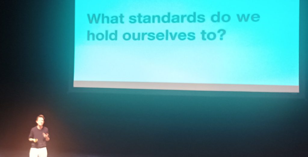 Lou Downe presenting on stage next to a slide reading 'What Standards do we hold ourselves to?'